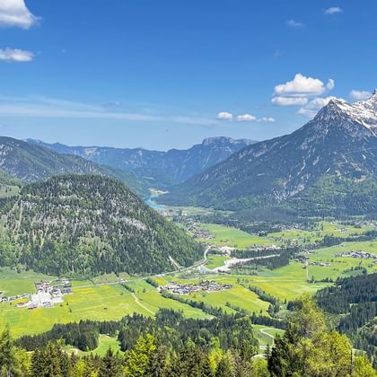 Panoramic view of a green Austrian alpine valley with villages, surrounded by forested mountains and snow-capped peaks under blue sky.