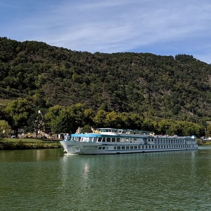 Hvidt flodkrydstogtskib Switzerland sejlende på roligt grønt vand med skovklædte bakker og lille landsby i baggrunden under blå himmel.