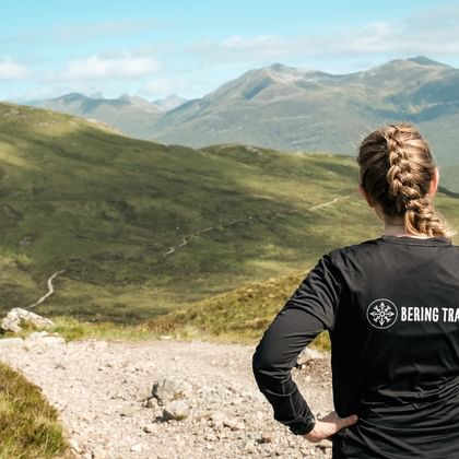 Kvinde med flettet hår i Bering Travel jakke står på sti med udsigt over grønne skotske højlande med bjerge under blå himmel.