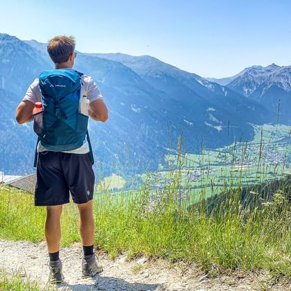Vandrare med blå ryggsäck på bergsled med utsikt över Alpdal med gröna ängar och snötäckta toppar vid Garmisch.