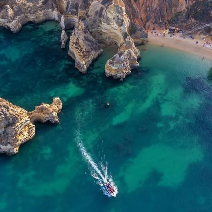 Aerial view of turquoise waters and golden cliffs in Lagos, Portugal. A small boat creates a white wake trail in the emerald sea near rock formations.