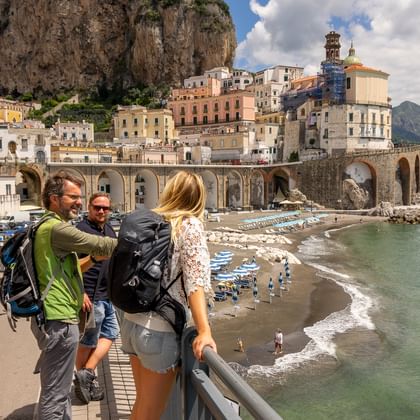 Tre turister med rygsække ved udsigtspunkt over kystby bygget ind i klipper med strand og turkis hav nedenfor.