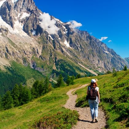 Ensom vandrer med rygsæk på jordsti gennem grønne alpenge med sne-dækkede bjergtoppe og blå himmel i Frankrig.