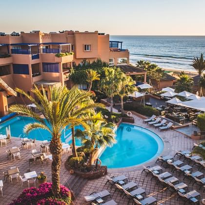 Aerial view of Paradise Plage resort in Agadir with curved swimming pools, palm trees, sun loungers, and beachfront location at sunset.