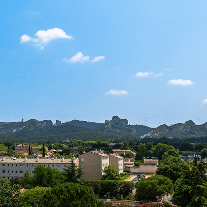 Panoramaudsigt over Saint-Rémy-de-Provence med terracottatage og cypresser, med de klippefyldte Alpilles-bjerge i baggrunden under blå himmel.