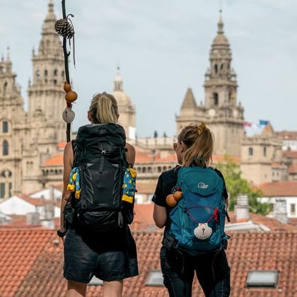 Two hikers with backpacks viewing Santiago de Compostela Cathedral from a rooftop. The cathedral's towers rise above red-tiled roofs.