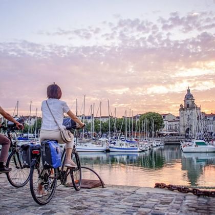 To cyklister med turcykler og tasker sidder ved solnedgang ved La Rochelles historiske havn og ser ud over både og klokkentårnet.