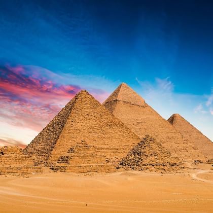 Tre gamle pyramider i Giza i den egyptiske ørken under en dramatisk solnedgangshimmel med orange og blå skyer.