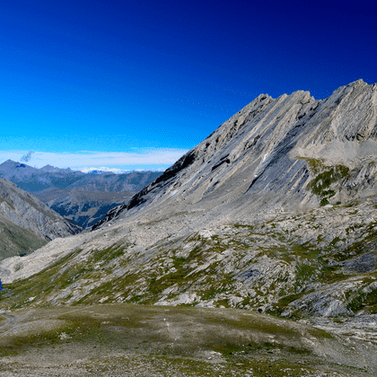 Klippefyldt bjergtop med lagdelte grålige skråninger over en lille blå alpesø i Queyras Naturpark, Frankrig, under klar blå himmel.