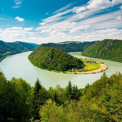 Luftfoto af Donau, der danner en hestesko-sløjfe omkring en skovklædt bakke i Østrig, med grønne skove og blå himmel.
