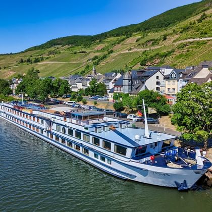 MS Olympia flodkrydstogtskib fortøjet ved en kaj ved siden af en tysk landsby med vingårdsdækkede bakker i baggrunden.