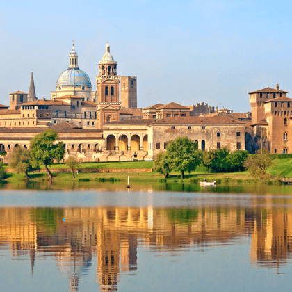 Historisk Mantova skyline med domkuppel og klokketårn spejlet i roligt Po-vand, omgivet af grøn vegetation under blå himmel.