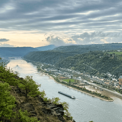 Panoramaudsigt over Rhinen, der slynger sig gennem dalen med fragtskib, byer på begge bredder, grønne bakker og dramatisk skyet himmel.
