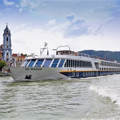 Hvidt flodkrydstogtskib MS SE-MANON sejlende på Donau foran historisk by med barok kirketårn og røde tage.