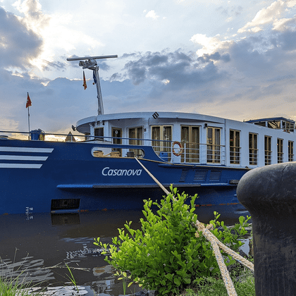 Blåt og hvidt flodkrydstogtskib MS Casanova fortøjet ved en grøn flodbred ved skumring. En pullert med reb er synlig i forgrunden.