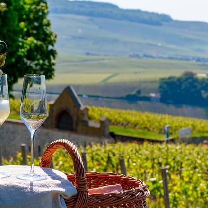Hånd heller champagne i glass på hvitt bord med kurv, med utsikt over Champagne-vingårder og bølgende åser.