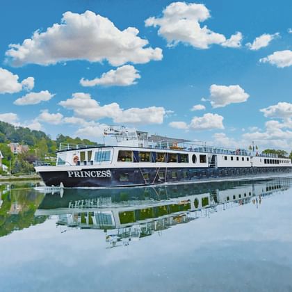 Hvidt flodkrydstogtskib MS Princess fortøjet på stille vand med perfekt reflektion. Blå himmel med hvide skyer og grønne bakker.