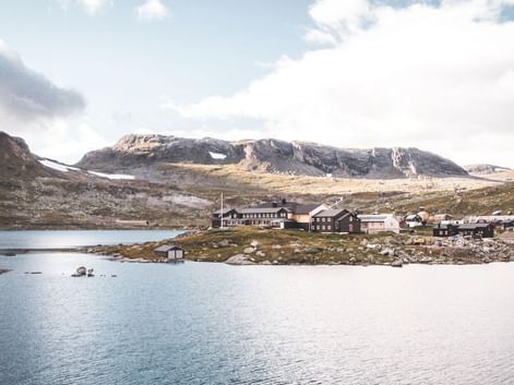 DNT Finsehytta hyttekompleks på Hardangervidda-plateauet ved en bjergsø med snedækkede toppe i baggrunden under skyet himmel.