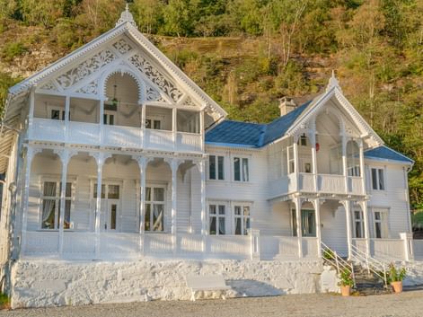 Hvidt to-etagers træhotel med udsmykkede detaljer og balkoner ved en grøn skråning i Norge.