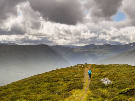 Vandrer i blå jakke på græsklædt bjergsti langs Kongevegen i Norge, med dramatisk overskyet himmel og tåget dal.