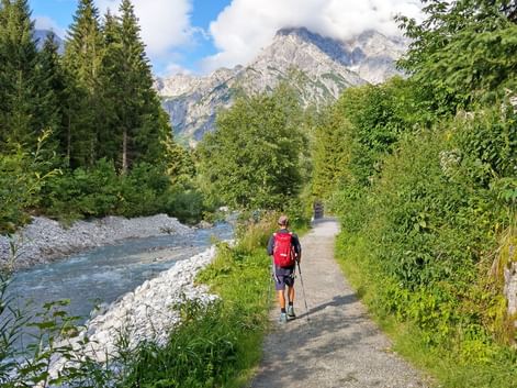 Hiker with red backpack walking on gravel path beside turquoise river in Austrian Alps, surrounded by green forest and rocky peaks.
