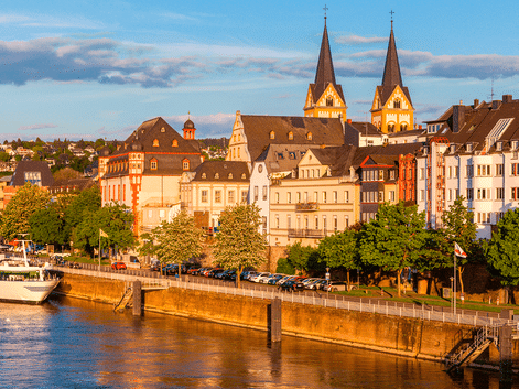 Aftenvisning af Koblenz flodfront med historiske bygninger og kirke med tvillingetårne. Et krydstogtskib er fortøjet langs promenaden.