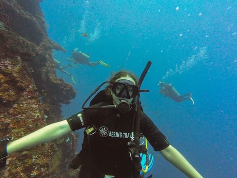 Undervandsselfie af dykker i sort våddragt med Bering Travel-logo, der udforsker koralrev i Balis klare blå vand med andre dykkere synlige.