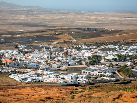Aerial view of Teguise, a white-walled town on Lanzarote island, surrounded by volcanic landscape with mountains in the background.
