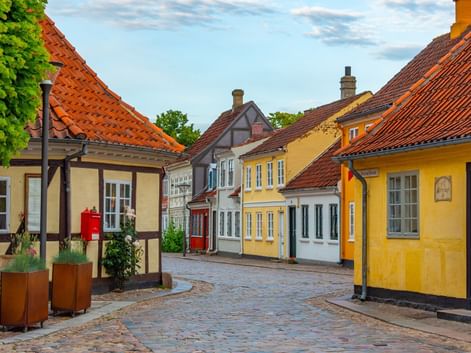 Kullerstensgata i Odense med färgglada korsvirkeshus, gula och vita fasader och röda tegeltak under blå himmel.