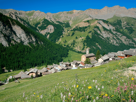 Landsby Saint-Véran med stenhuse og kirketårn på en skråning. Vildblomstereng i forgrunden, skovklædte bjerge bagved.