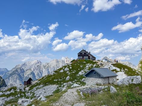 Alpina stugor på en klippig bergskam i Juliska Alperna med taggiga bergstoppar i bakgrunden under en blå himmel med vita moln.