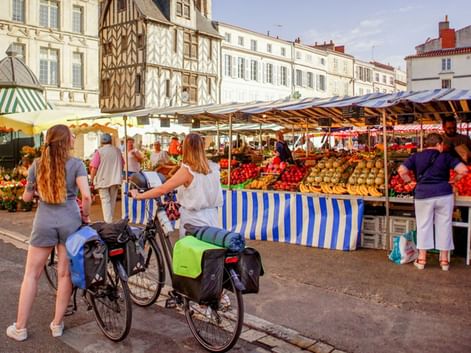 Udendørs marked i La Rochelle med frugt- og grøntsagsboder. To cyklister med cykeltasker ser på markedet foran historiske bygninger.