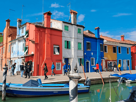 Række af farverige huse i rødt, hvidt, grønt, blåt og orange langs en kanal med fortøjede både i Venedig under blå himmel.