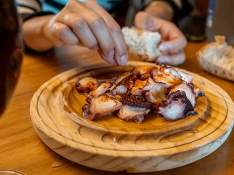 Grillede blekkspruttentakler servert på en rund tretallerken. Hender som holder brød er synlige i bakgrunnen på et trebord.