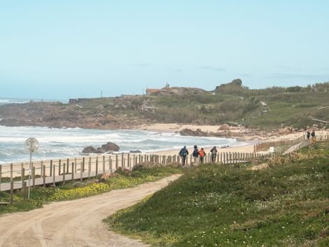 Pilgrims walking on sandy path with wooden fence along Atlantic coast on Portuguese Camino. Green vegetation and beach visible with ocean waves.