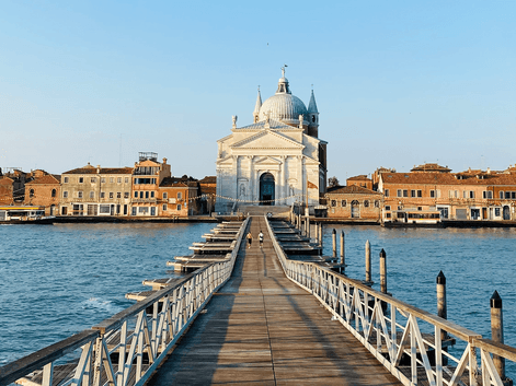 Lang træbro over blåt vand til hvid kuppelkirke i Venedig. Historiske bygninger langs vandkanten under klar himmel.