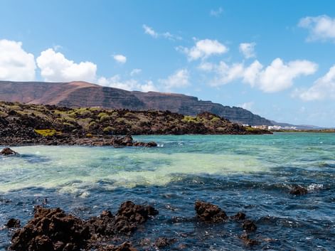Volcanic coastline near Orzola, Lanzarote with turquoise water, black lava rocks, and barren hills under a blue sky with white clouds.