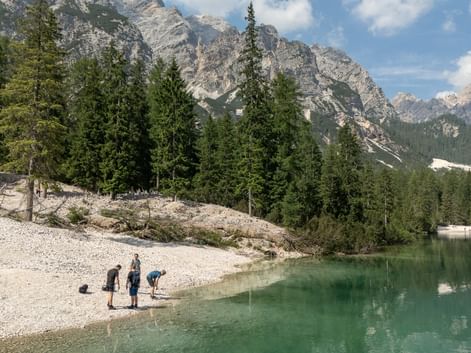 Vandrere ved Dolomitternes bjergsø Tre vandrere på en stenstrand ved en turkis bjergsø i Dolomitterne, med dramatiske kalkstenstinder og nåleskov.