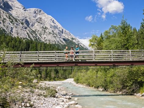 To vandrere på en træbro over en turkis bjergbæk, med klippefyldt Zugspitze-top og grøn skov i baggrunden.