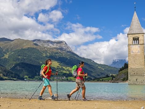 Två vandrare med ryggsäckar och vandringsstavar längs stranden av Reschensee med ett historiskt klocktorn i vattnet och berg i bakgrunden.