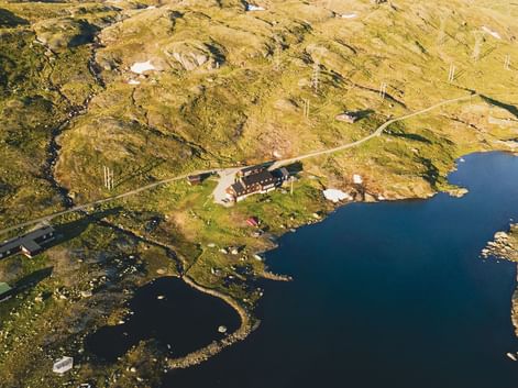 Luftfoto af Geiterygghytta DNT-hytte på Hardangervidda, beliggende på en halvø mellem blå bjergsøer omgivet af gylden tundra.