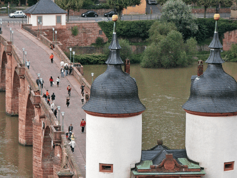Heidelbergs gamle bro over Neckar med fodgængere. To hvide baroktårne med grønne kupler indramme broindgangen.