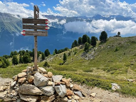 Träskylt på stenkummel i alpin äng med bergstoppar och moln i bakgrunden på vandringsleden Garmisch till Merano.