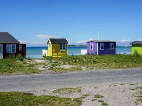 Fyra färgglada strandstugor i rött, gult, lila och grönt längs en sandstrand på ön Ærø med blått hav och klar himmel i bakgrunden.