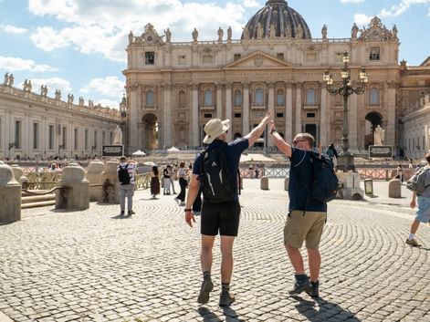 Two hikers with backpacks raising arms in celebration on St. Peter's Square in Rome, with St. Peter's Basilica and colonnade in background.