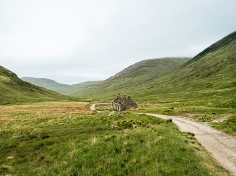 Grusvej der fører til stenruin i grøn dal på West Highland Way, Skotland. Bølgende bakker indramme dalen under overskyet himmel.