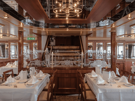 Elegant skibsrestaurant med hvide duge, træpaneler og central trappe. Reflekterende loft og panoramavinduer langs væggene.
