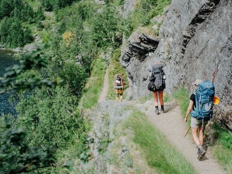 Tre vandrere med rygsække på en smal bjergsti i Aurlandsdalen, Norge. Klippevæg til højre, grøn dal med flod nedenfor.