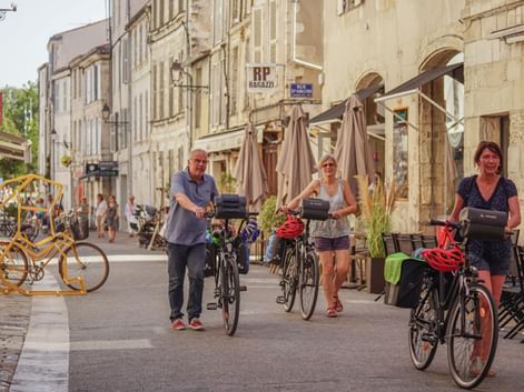 Tre cyklister med cykler går gennem en gågade i historiske La Rochelle, Frankrig. Stenbygninger med caféer langs begge sider.