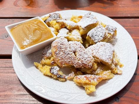 Austrian Kaiserschmarrn dusted with powdered sugar on a white plate, served with applesauce in a small bowl on a rustic wooden table.